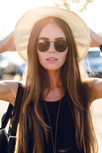 Free photo stylish girl standing near road wearing short black dress, straw hat, black eyeglasses, and black backpack. she smiles in the warm rays of setting sun. she touches her sunglasses with hand