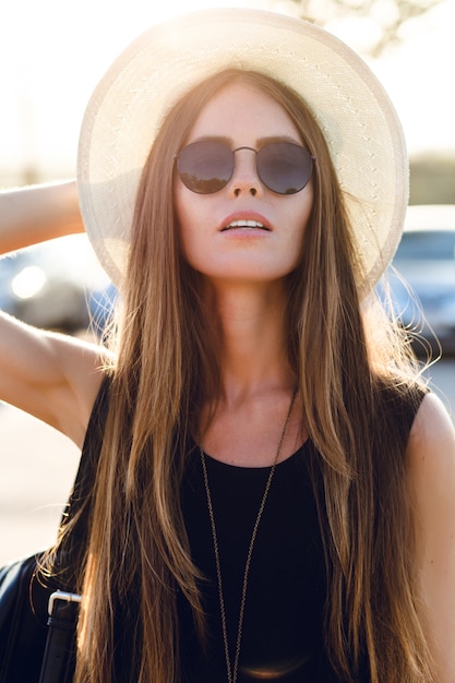 Stylish girl standing near road wearing short black dress, straw hat, black eyeglasses, and black backpack. She smiles in the warm rays of setting sun. She touches her sunglasses with hand