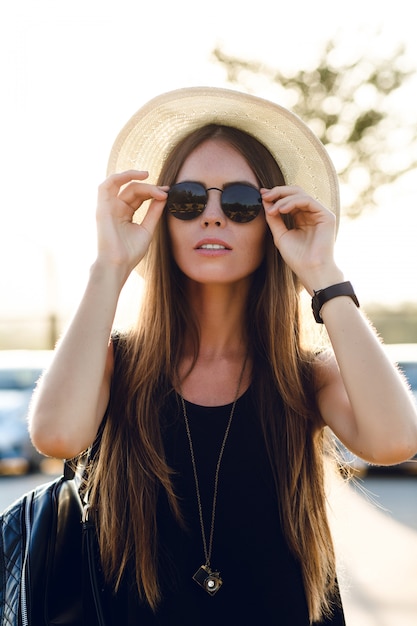 Stylish girl standing near road wearing short black dress, straw hat, black eyeglasses, and black backpack. She smiles in the warm rays of setting sun. She touches her sunglasses with hand