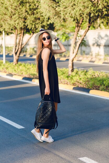 Stylish girl standing on the road wearing short black dress, straw hat, black eyeglasses, white sneakers and holding black backpack. She smiles in the warm rays of setting sun