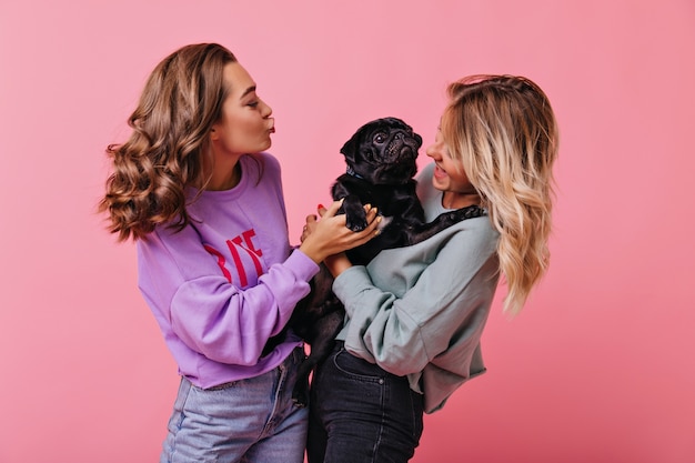 Stylish girl with brown shiny hair looking at dog with kissing face expression. Indoor portrait of cheerful blonde woman standing on rosy with her puppy.