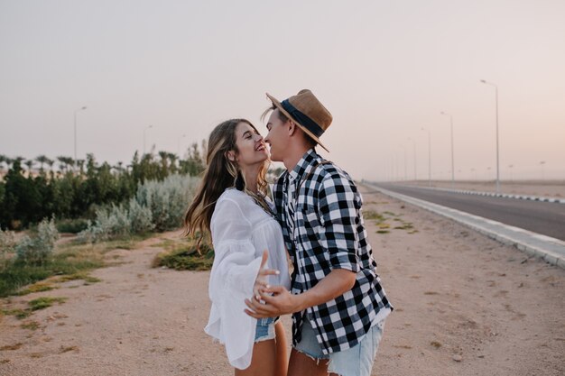 Stylish guy and adorable woman are dancing on the sidelines, enjoying a warm evening and smiling. Laughing couple in trendy outfit gently hugging, walking along the road at sunset and having fun