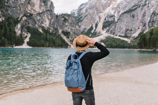 Free photo stylish guy wearing vintage hat decorated with ribbon relaxing on the lake shore and looking at water