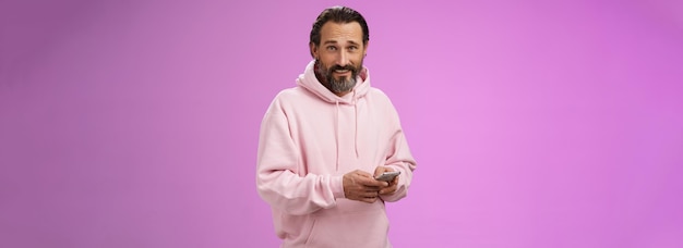 Free photo stylish handsome adult bearded guy s in trendy pink hoodie holding smartphone texting look camera un