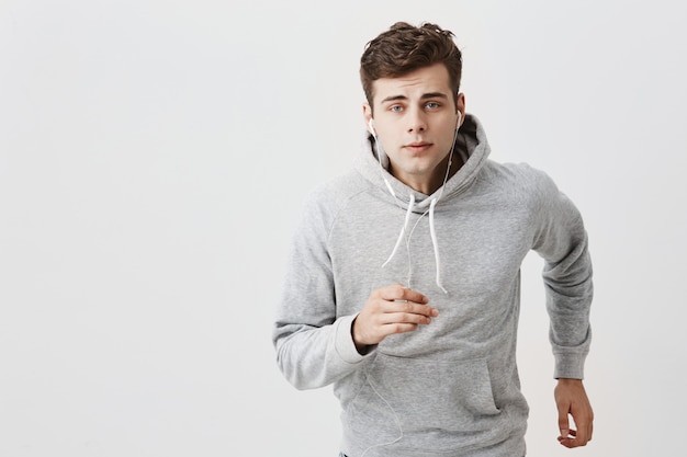 Free photo stylish handsome european young man with trendy haircut dressed in gray hoodie and jeans runs, looks straight forward with blue eyes, listens to favourite songs via white earphones, music app.