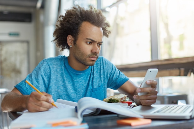 Stylish handsome guy with dark skin sitting at cafeteria writing something in his textbook holding cell phone browsing newsfeed via social networks online looking puzzled finding out bad news