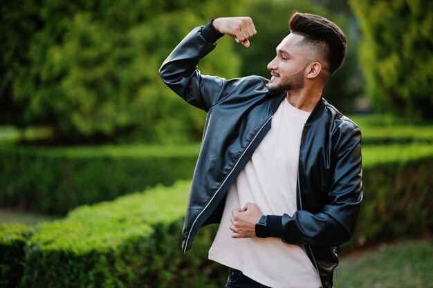 Stylish indian beard man at black leather jacket shows his muscle hand India model posed at green garden of city