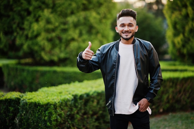 Stylish indian beard man at black leather jacket shows thumb up India model posed at green garden of city