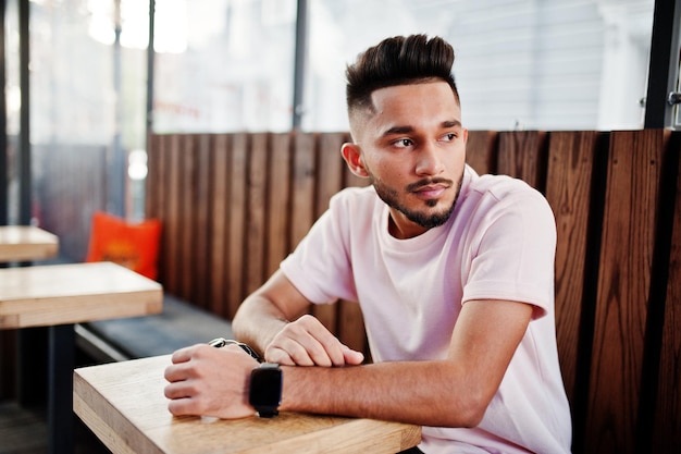 Stylish indian beard man at pink tshirt sitting against wooden table of cafe India model posed outdoor