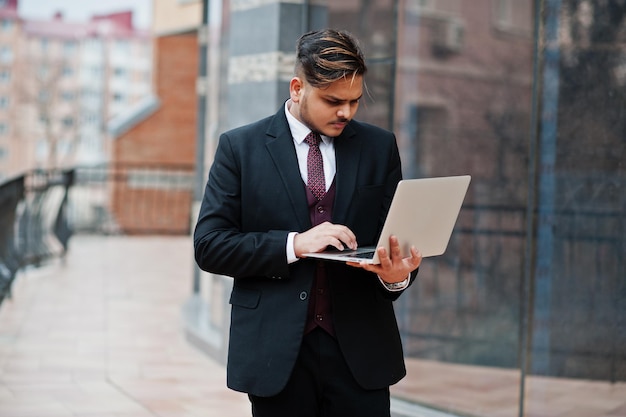 Free photo stylish indian businessman in formal wear with laptop on hands standing against windows in business center