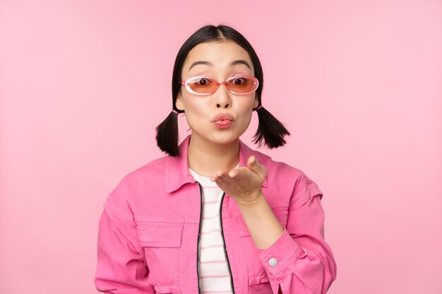 Stylish kpop girl in sunglasses asian woman blowing air kiss at camera pucker lips mwah gesture standing over pink studio background