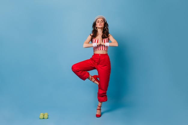 Free Photo stylish lady in red pants and striped cropped top standing in tree pose on blue wall with green dumbbells