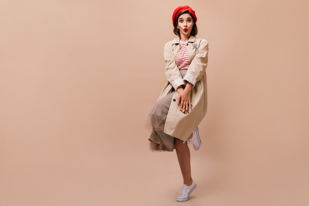 Free Photo stylish lady in trench and hat shyly poses on beige background.  cute girl in striped sweater, in beige cloak and red beret jumps.