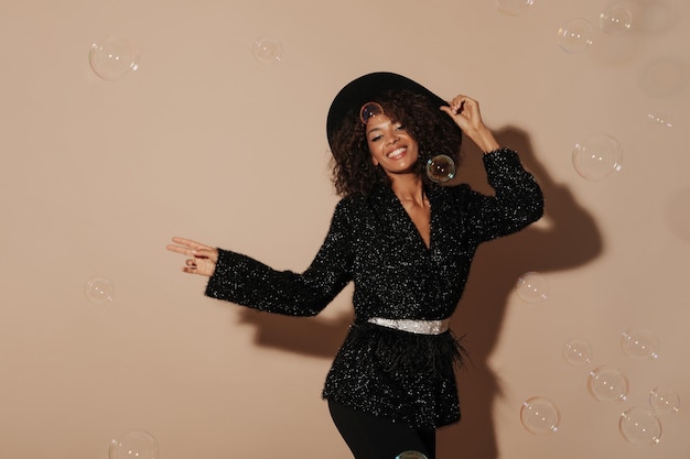 Stylish lady with brunette hairstyle in shiny sweater with light belt and black hat smiling and looking into camera on beige backdrop