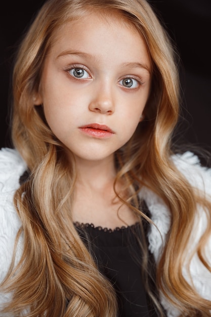 Stylish little  girl posing in white outfit isolated on black  wall. Beautiful caucasian blonde female model's posing. Human emotions, facial expression, childhood, fashion, style.