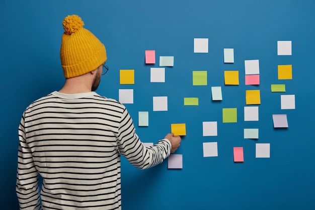 Free Photo stylish male youngster in fashionable outfit stands back to camera, turns to blue wall, sticks small colorful notes for putting down main ideas