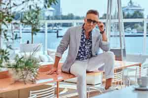 Free photo stylish man dressed in modern elegant clothes correct his sunglasses while sitting on a table at outdoor cafe against the background of city wharf.