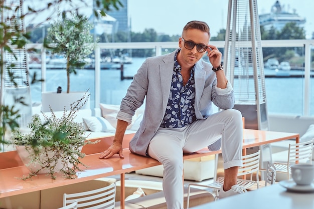 Stylish man dressed in modern elegant clothes correct his sunglasses while sitting on a table at outdoor cafe against the background of city wharf.