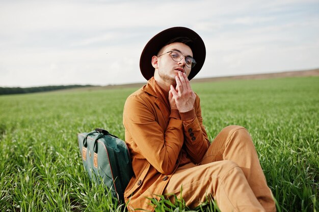 Stylish man in glasses brown jacket and hat with bag posed on green field