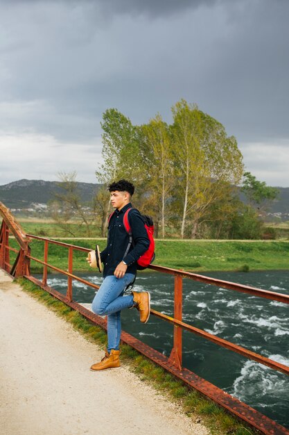 Stylish man holding hat leaning on bridge railing over flowing river