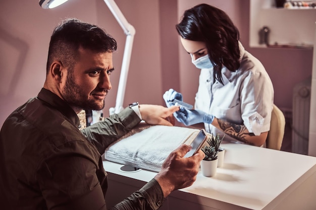 A stylish man in shirt holding a phone and looking sideways while receiving a manicure by a beautician in the beauty salon