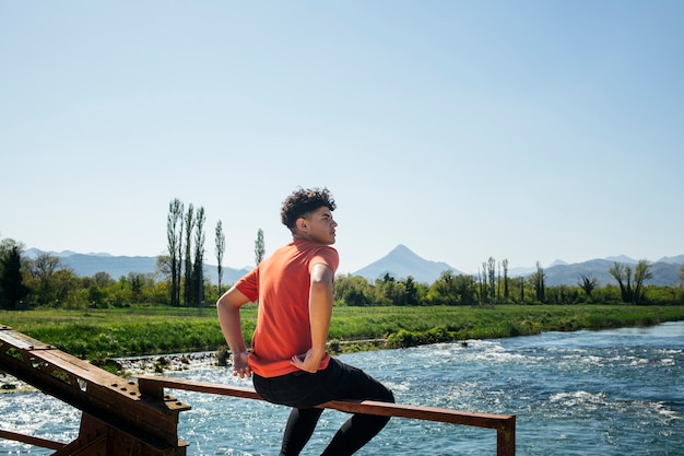 Stylish man sitting on metallic railing of bridge over river