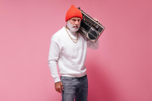 Stylish man in white sweater posing with record player Serious bearded guy in orange hat with gold chains looking into camera