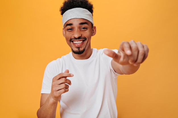 Free photo stylish man in white tshirt laughs and points his finger at camera portrait of brunette guy in white headband smiling and dancing on orange background