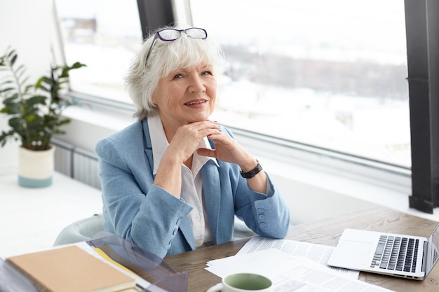 Stylish mature middle aged woman writer with gray hair and wrinkles looking and smiling happily, clasping hands, being in good mood, feeling inspired while working on her new book