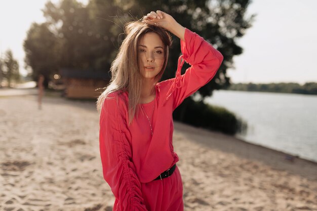 Stylish model with flying dark haired dressed pink summer dress is looking at camera and touching her hair on background of sandy beach in sunshine