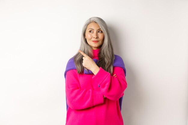 Stylish modern asian elderly woman pointing at upper left corner, looking at logo with pleased smile, standing in pink sweater over white background