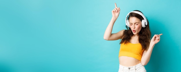 Free Photo stylish modern woman in headphones listening music and dancing enjoying favorite songs in earphones standing against blue background