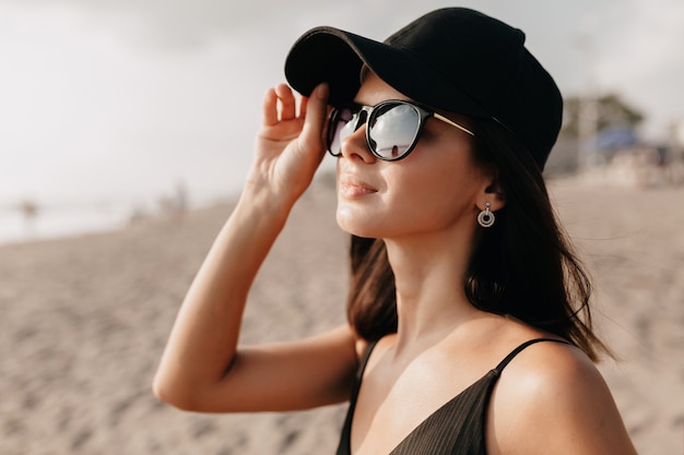 Stylish modern woman in trend outfit looking on the ocean with happy smile wearing cap and glasses and enjoys summer hot days  Young caucasian female model on the sea shore