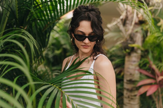 Stylish modern young woman with blond curly hairstyle wearing summer clothes walking among tropics in summer sunny day. Outside photo of happy smiling girl has fun and enjoying weekend