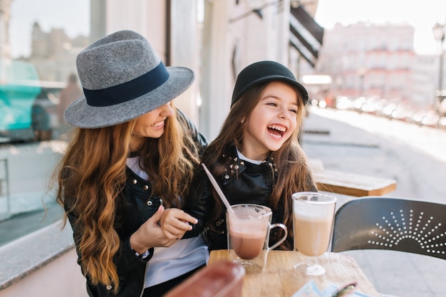 Stylish mom and pretty smiling daughter enjoying weekend together in outdoor restaurant drinking coffee and milk shake.