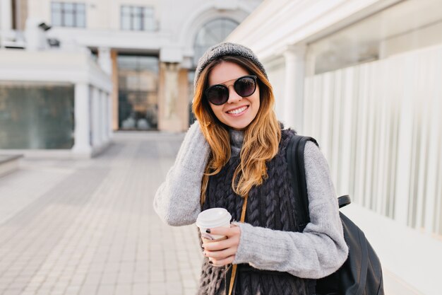 Stylish portrait joyful fashionable woman with long blonde hair, knitted hat, warm woollen sweater and modern sunglasses walking in city centre. Travelling with coffee to go, happiness. Place for text.