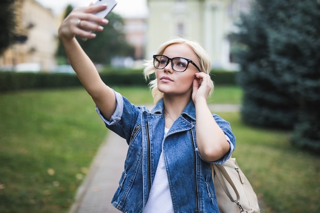 Stylish pretty fashion blonde girl woman in jeans suite makes selfie on her phone in the city in the morning