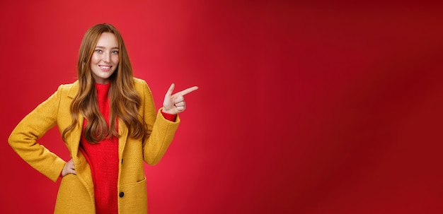 Free photo stylish and selfassured attractive redhead woman with freckles in yellow outdoor coat pointing right