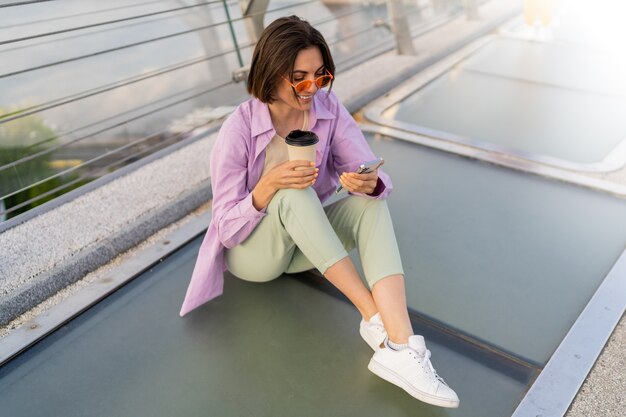 Stylish short haired woman sitting on modern bridge, enjoying coffee and using mobile phone