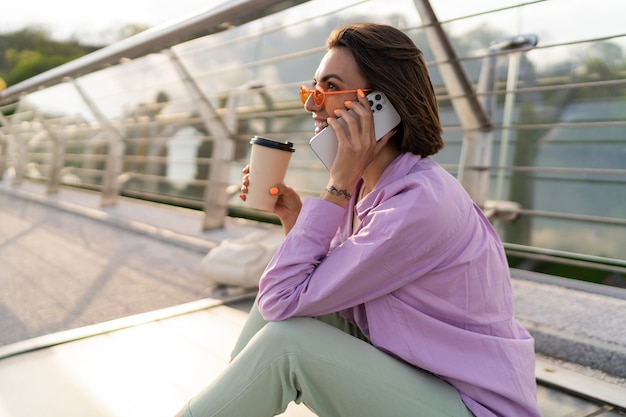 Stylish short haired woman sitting on modern bridge, enjoying coffee and using mobile phone