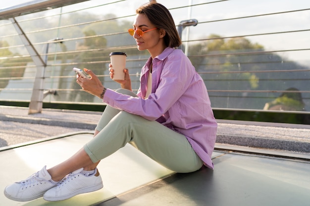 Stylish short haired woman sitting on modern bridge, enjoying coffee and using mobile phone