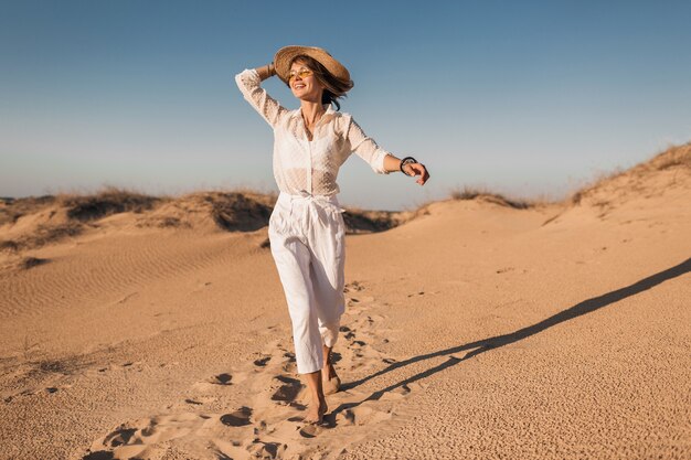 Stylish smiling beautiful happy woman running and jumping in desert sand in white outfit wearing straw hat on sunset