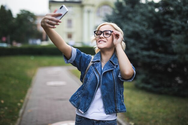 Stylish smiling fashion blonde girl woman in jeans suite makes selfie on her phone in the city in the morning