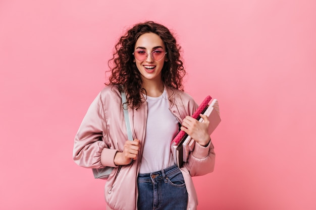 Stylish student in sunglasses poses with notebooks on pink background