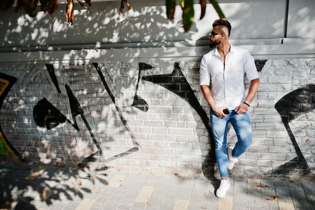 Free Photo stylish tall arabian man model in white shirt jeans and sunglasses posed at street of city beard attractive arab guy against graffiti wall