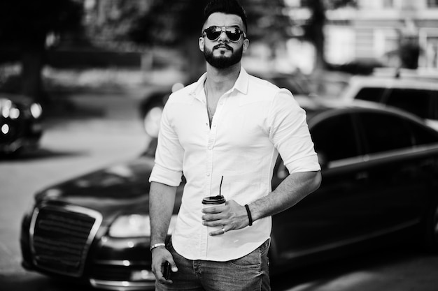 Free photo stylish tall arabian man model in white shirt jeans and sunglasses posed at street of city beard attractive arab guy with cup of coffee against black business car