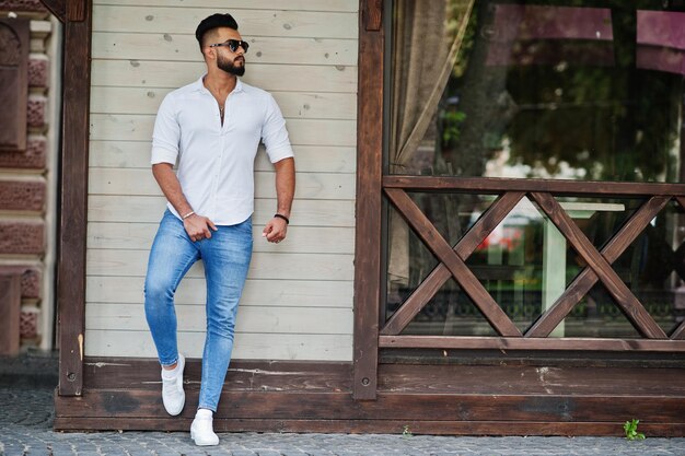 Stylish tall arabian man model in white shirt jeans and sunglasses posed at street of city Beard attractive arab guy