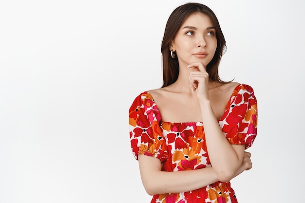 Stylish thoughtful girl in feminine floral dress looking at upper right corner and thinking making choice standing over white background