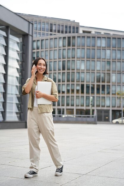 Stylish urban girl walking on street talking on mobile phone and carrying laptop young woman making