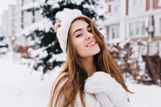 Free photo stylish winter portrait of charming young woman with long brunette hair, in woollen white gloves and hat walking on street full with snow. cheerful mood, smiling.